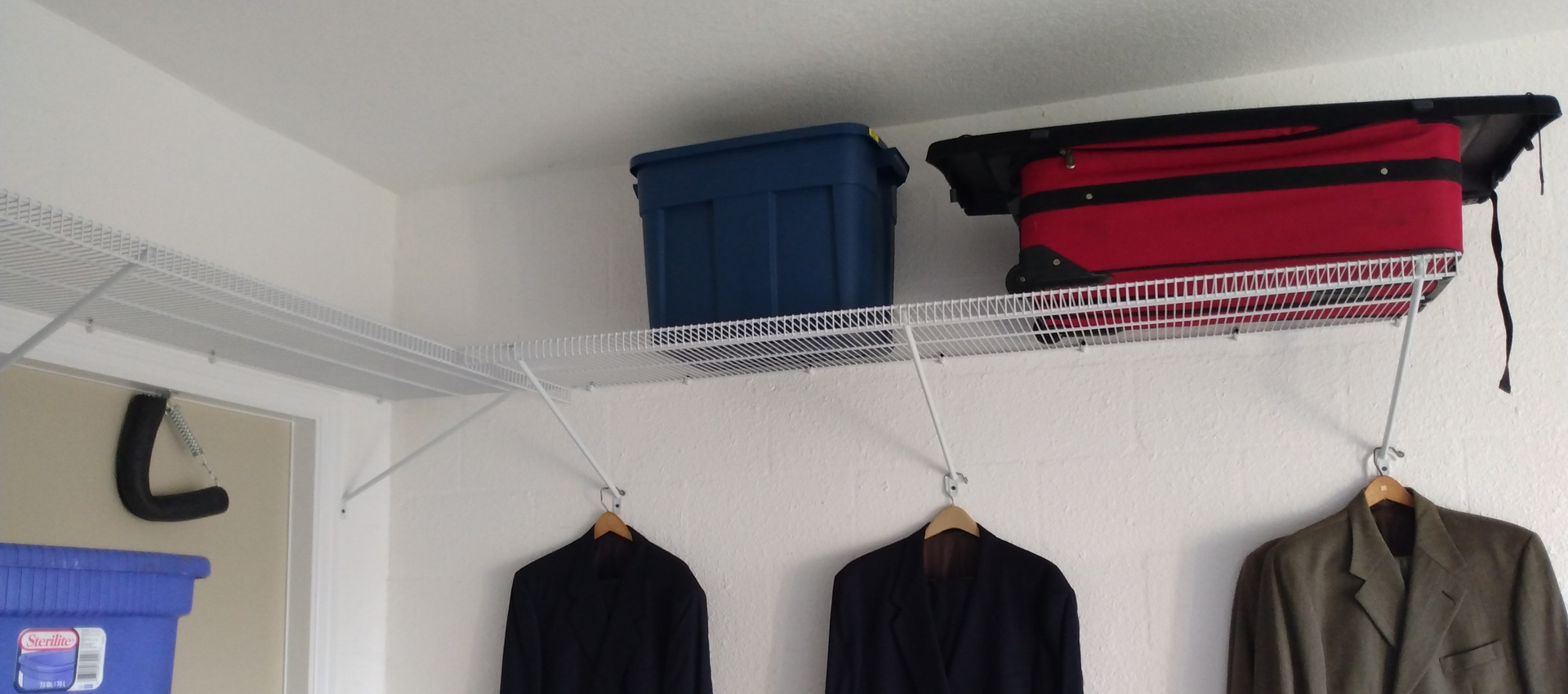 White metal wire shelves ClosetMaid installed on masonry block wall. Plastic boxes and suits on it.