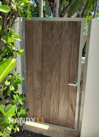 White concreete fence, brown wood entrance door before repair, green plants around.