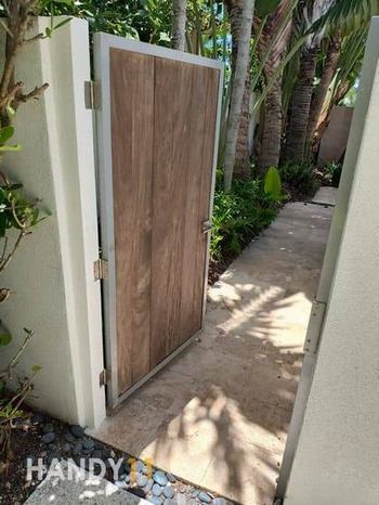 White concreete fence and opened entrance brown door. Green plants around.