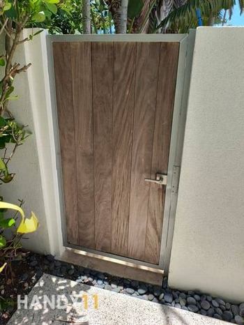 Brown wood and grey metal frame entrance door repaired. White concreete fence. Green plants around.