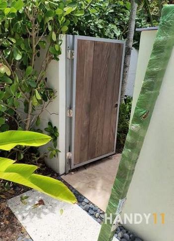 IPE wood post in green film. White concreete fence, brown wood entrance door to repair, green plants around.